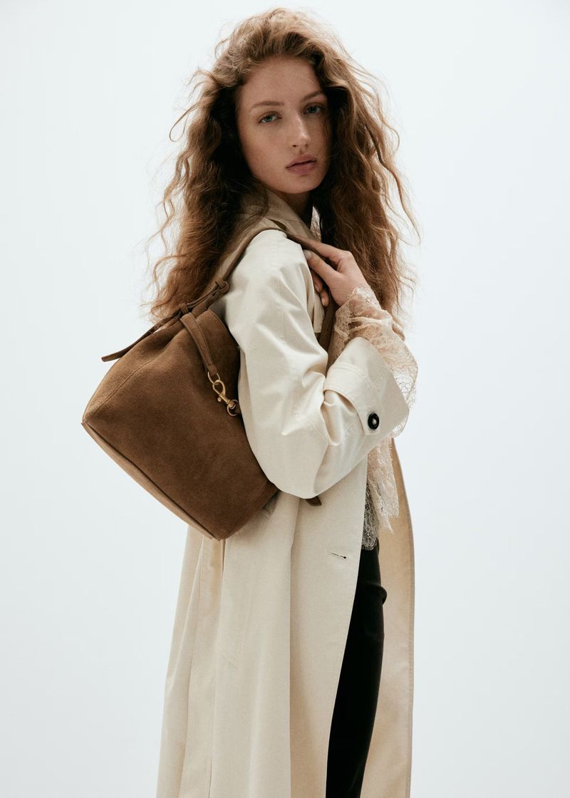Model with long curly hair wearing an off-white coat and holding a brown suede shoulder bag against a white background