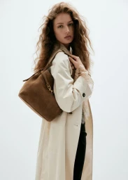 Model with long curly hair wearing an off-white coat and holding a brown suede shoulder bag against a white background