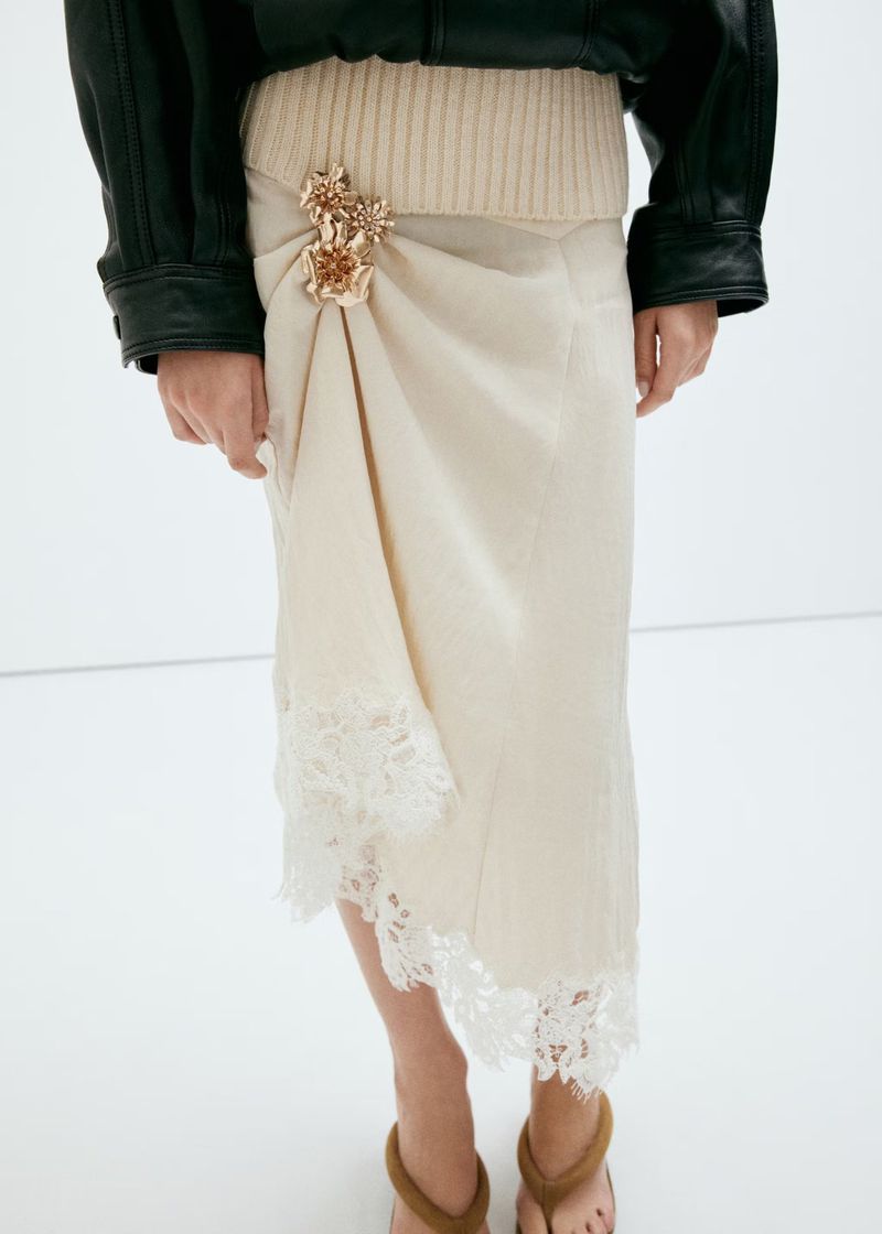 Model wearing a cream skirt with lace trim, a ribbed knit waist, and gold floral brooch, styled with a black jacket