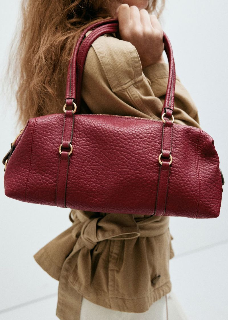 Model holding a textured red leather handbag wearing a beige utility jacket against a plain white background