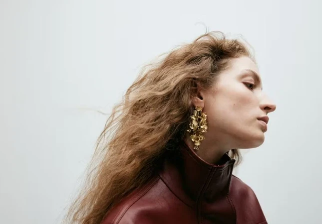 Profile portrait of a model with long curly hair wearing ornate gold floral earrings and a dark burgundy leather jacket against a white background.