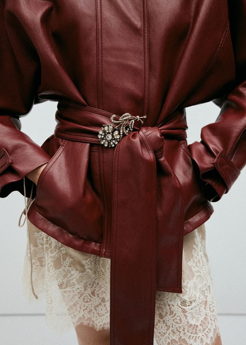 Model wearing a deep red leather jacket with a belted waist and floral brooch over a cream lace skirt