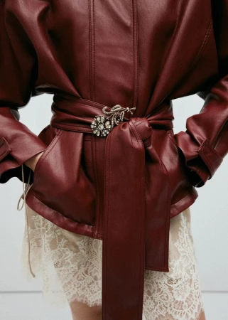 Model wearing a deep red leather jacket with a belted waist and floral brooch over a cream lace skirt