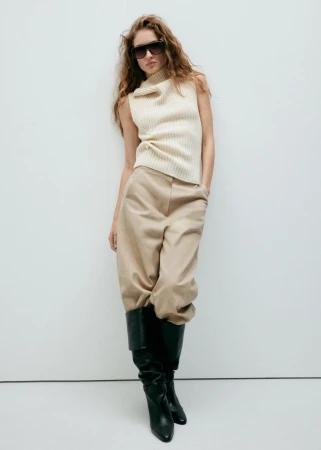 Model in dark sunglasses wearing a cream knit sleeveless top and beige trousers against a bright white background