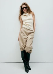 Model in dark sunglasses wearing a cream knit sleeveless top and beige trousers against a bright white background