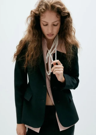 Model with long wavy hair wearing a black blazer and pearl necklace against a plain white background for Mango