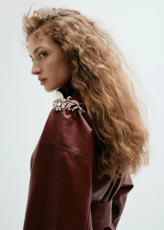 Model with long curly hair wearing a burgundy coat and rhinestone brooch against a white background