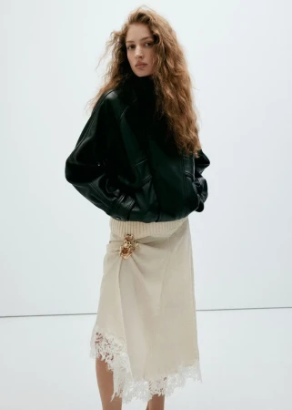 Model with long curly hair wearing a black leather jacket and cream lace skirt against a plain white background