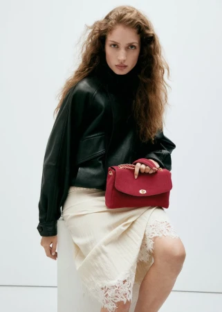 Model with long curly hair wearing a black leather jacket and white lace skirt holding a red handbag