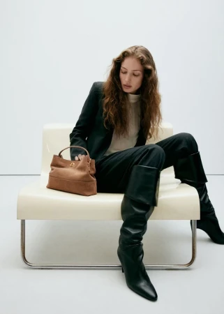 Model seated on a white bench wearing a dark blazer, trousers, and tall black boots, reaching for a brown handbag.