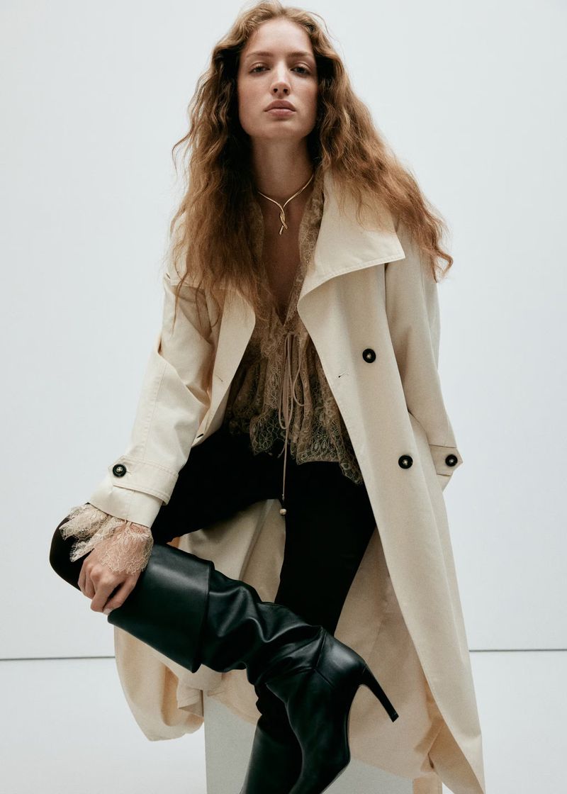 Model in cream trench coat and lace top with black boots against a white studio background