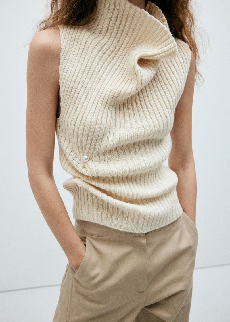 Model wearing a ribbed cream high neck sleeveless top and beige trousers against a plain white background