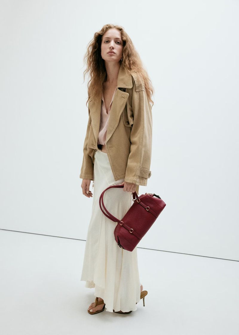 Model wearing a tan jacket and cream skirt holding a burgundy handbag in a studio