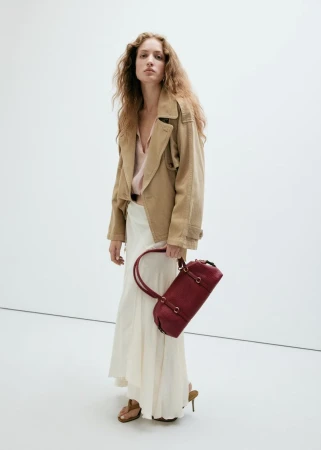 Model wearing a tan jacket and cream skirt holding a burgundy handbag in a studio