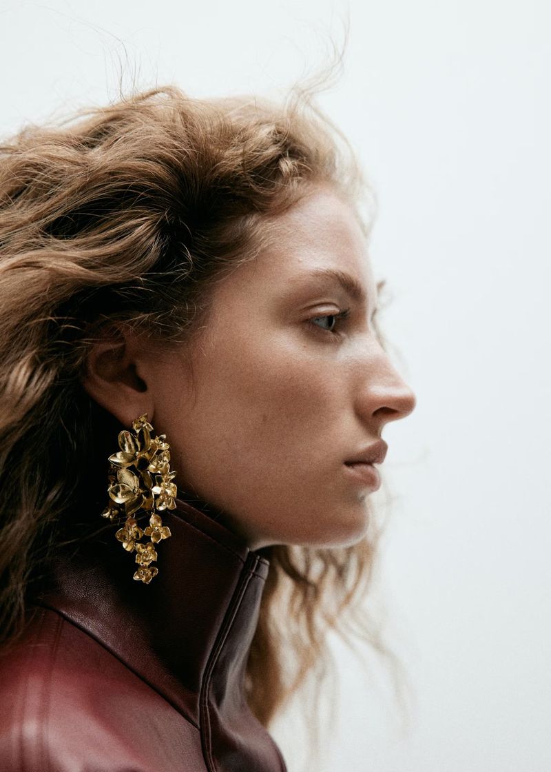 Profile portrait of a woman wearing statement gold floral earrings and a high-collared burgundy leather jacket against a light background.