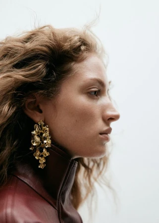 Profile portrait of a woman wearing statement gold floral earrings and a high-collared burgundy leather jacket against a light background.