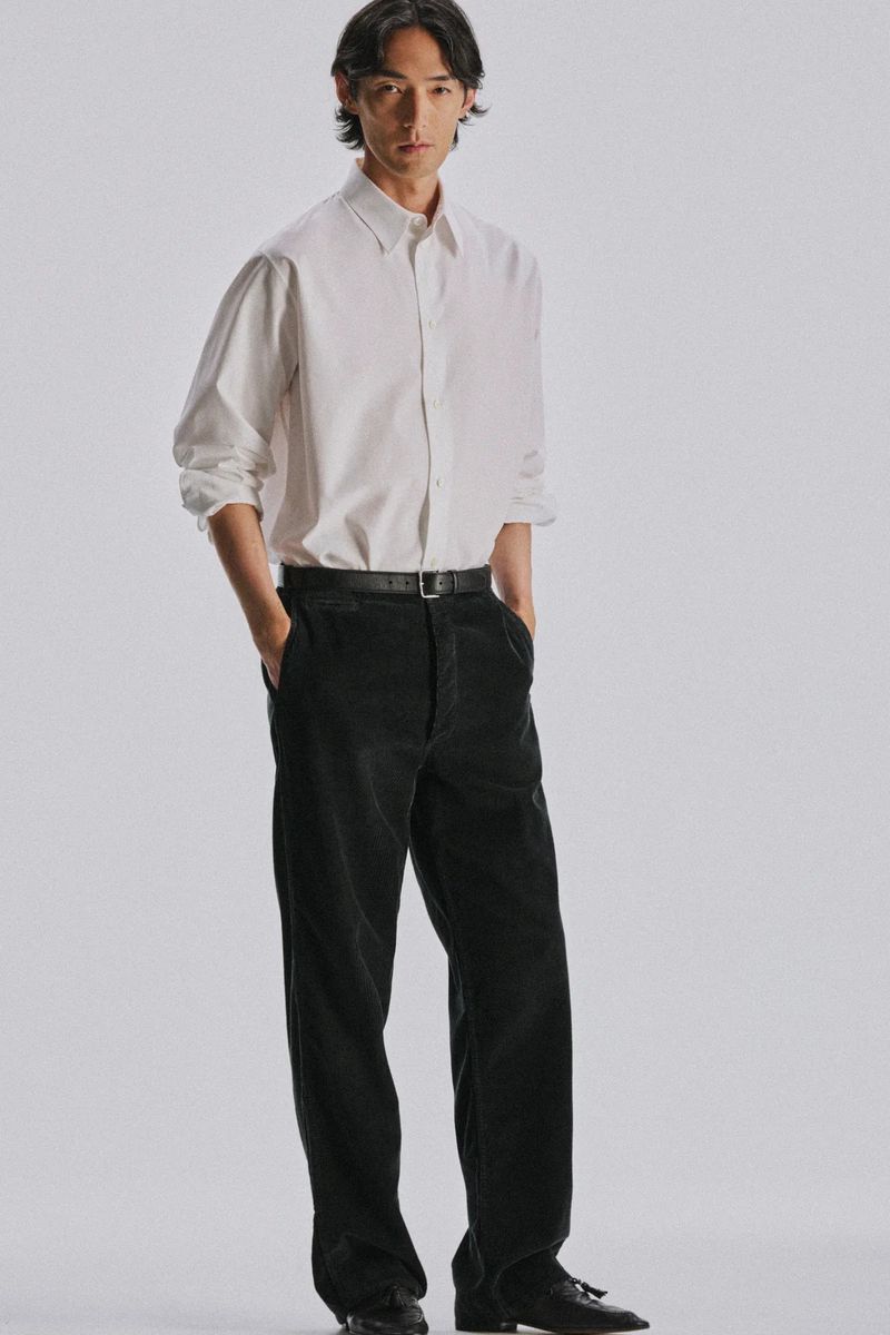 Man wearing a crisp white button-down shirt and black trousers standing against a pale grey seamless studio backdrop.