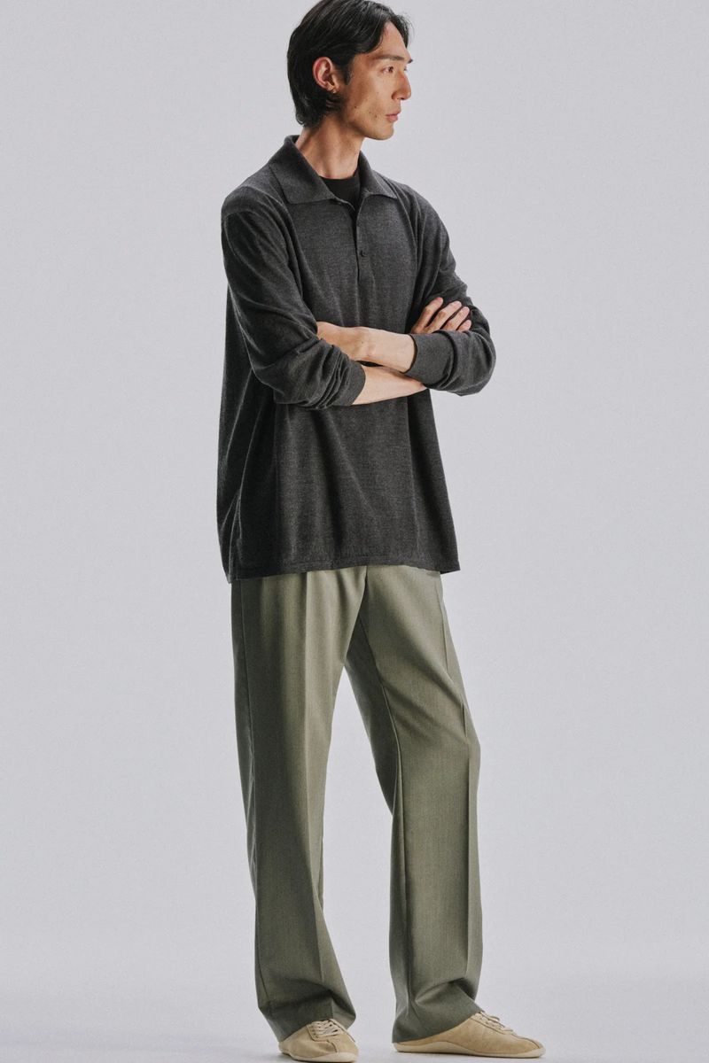 Model standing with arms crossed wearing a dark long-sleeve polo shirt and trousers against a grey studio backdrop.