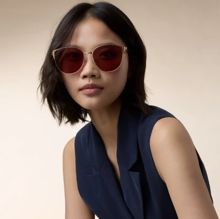 Model wearing gold-framed sunglasses with dark red lenses and a sleeveless navy blue top against a pale background