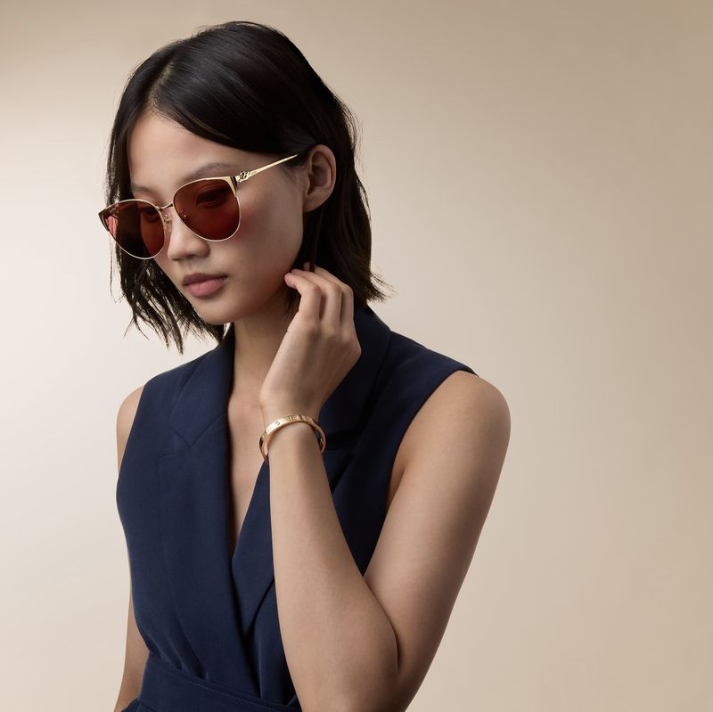 Model wearing gold sunglasses and a gold bracelet with a navy sleeveless vest top against a neutral background.