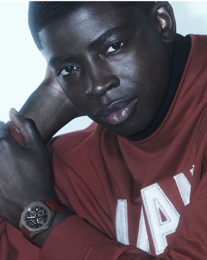 Portrait of a Black man wearing a red sweatshirt and wrist watch against a bright white background.
