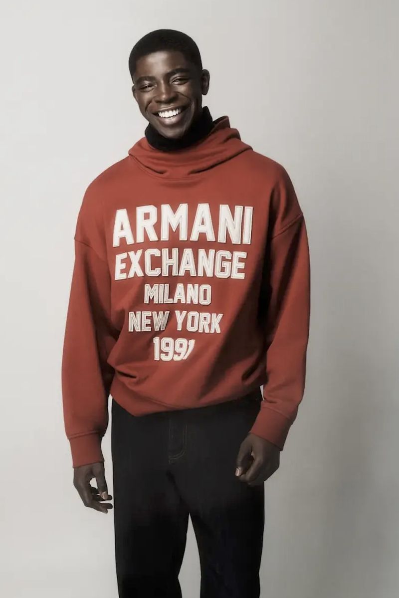 Smiling black man wearing a rust orange hooded sweatshirt with white lettering and dark pants against a plain grey background