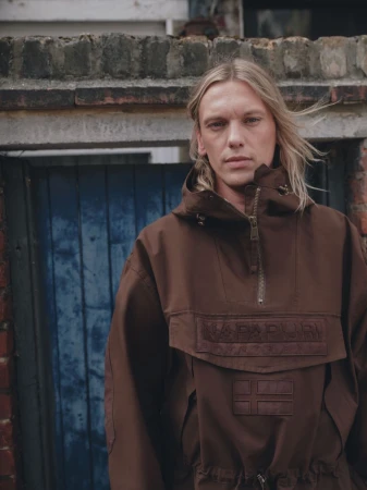 Jamie Campbell Bower wearing a brown jacket with a hood outside against a weathered brick and blue door background.