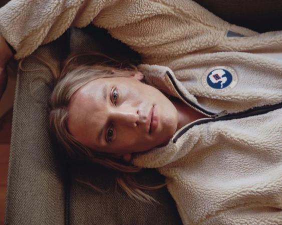 Jamie Campbell Bower in a cream fleece jacket reclining against a dark textured cushion