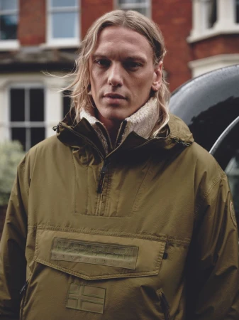 Jamie Campbell Bower wearing an olive green Napapijri hooded anorak jacket in front of a brick building exterior