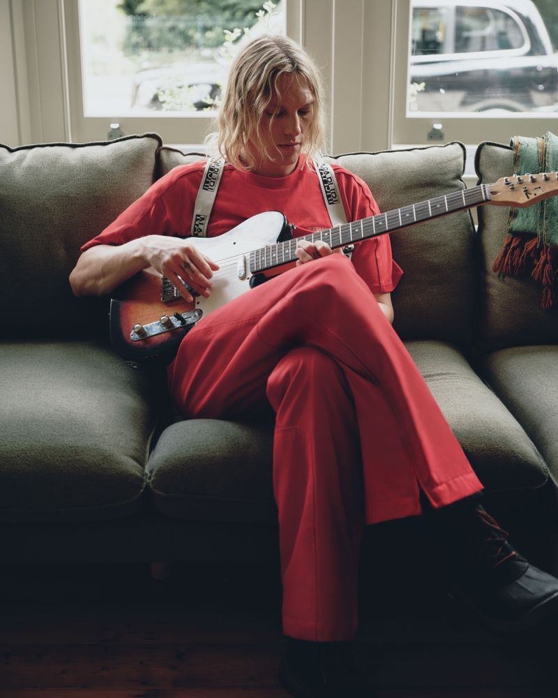Jamie Campbell Bower playing an electric guitar seated on a green sofa wearing a red outfit for the Napapijri FW26 campaign.