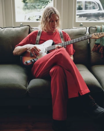 Jamie Campbell Bower playing an electric guitar seated on a green sofa wearing a red outfit for the Napapijri FW26 campaign.