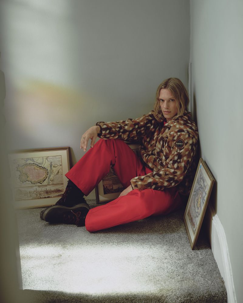 Jamie Campbell Bower seated in a corner wearing a patterned fleece jacket and red trousers for the Napapijri FW26 campaign.