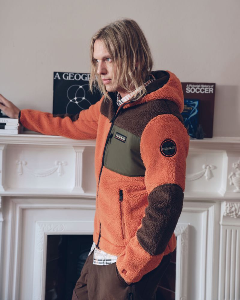 Jamie Campbell Bower wearing an orange fleece hooded jacket with brown and green panels standing indoors against a mantelpiece