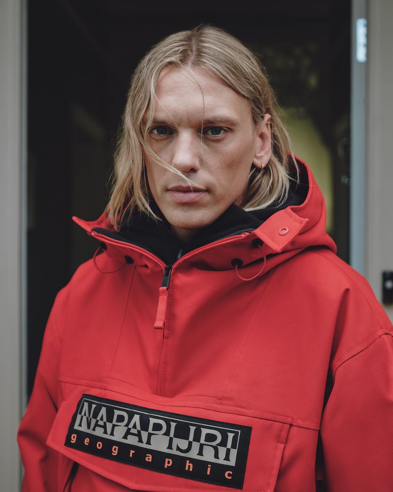 Jamie Campbell Bower in a bright red Napapijri hooded anorak jacket against a neutral doorway background