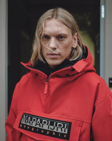 Jamie Campbell Bower in a bright red Napapijri hooded anorak jacket against a neutral doorway background