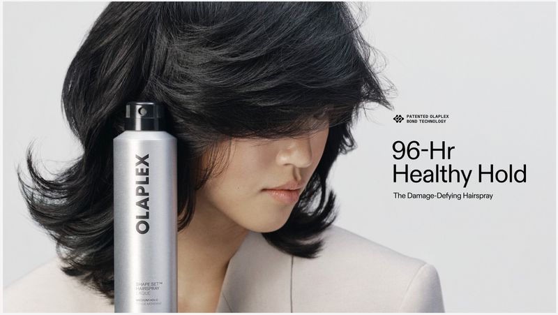 Close-up of a person with dark layered hair beside an Olaplex hairspray bottle against a light background with promotional text