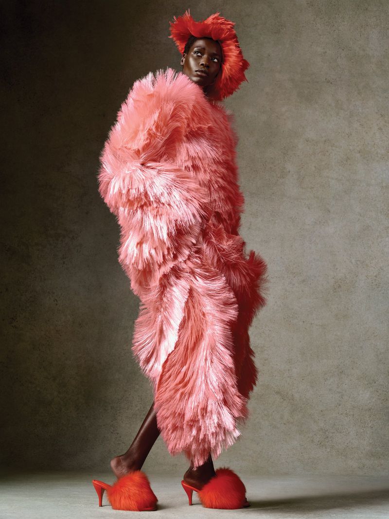 Model wearing a vibrant red feathered textured dress and matching mules against a neutral textured studio background.