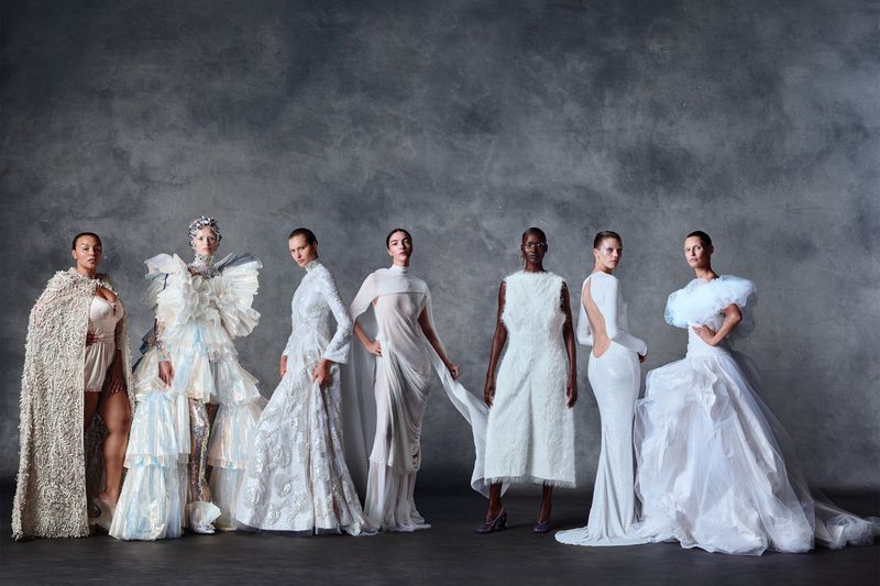 Group of seven female models wearing elaborate white and neutral fashion garments against a textured grey backdrop.