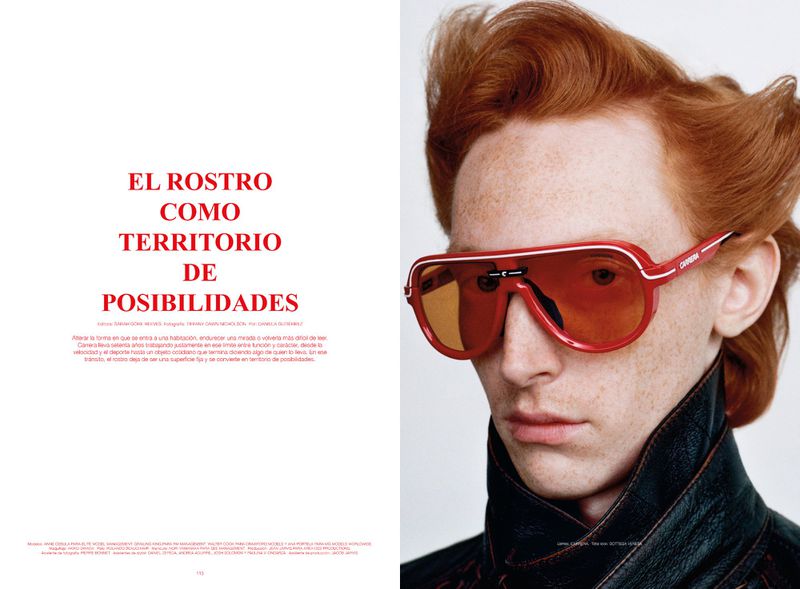 Close-up portrait of a red-haired model wearing statement red aviator sunglasses and a black leather jacket against a plain white background.