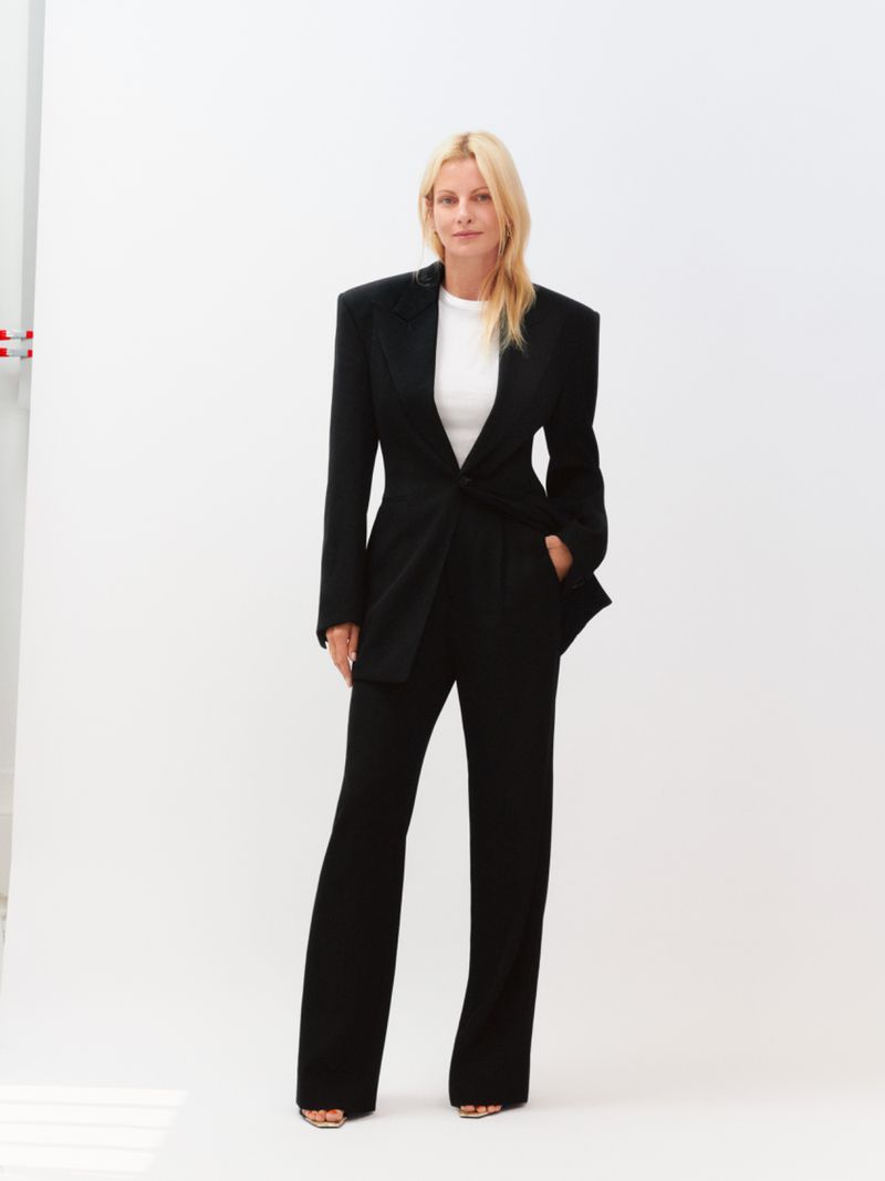 Blonde woman wearing a black tailored suit and white t-shirt standing against a white seamless backdrop.