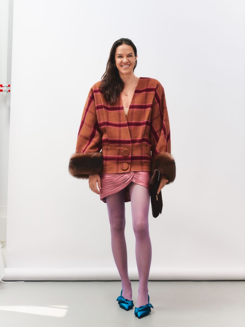 Woman wearing a brown plaid jacket with fur cuffs, pink miniskirt, purple tights, and blue heels against a white backdrop.