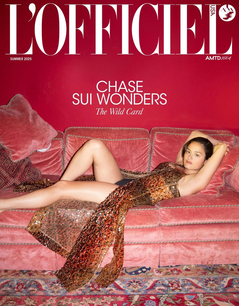 Chase Sui Wonders reclining across a pink velvet couch wearing a shimmering sequined bronze gown in front of a bold red wall.