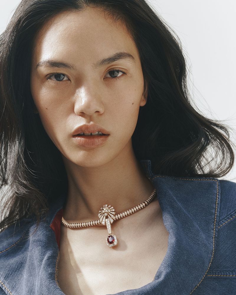 Close-up portrait of an East Asian model wearing a diamond and ruby necklace and denim jacket against a white backdrop.