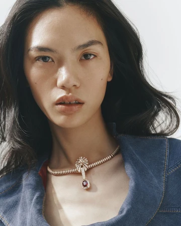 Close-up portrait of an East Asian model wearing a diamond and ruby necklace and denim jacket against a white backdrop.