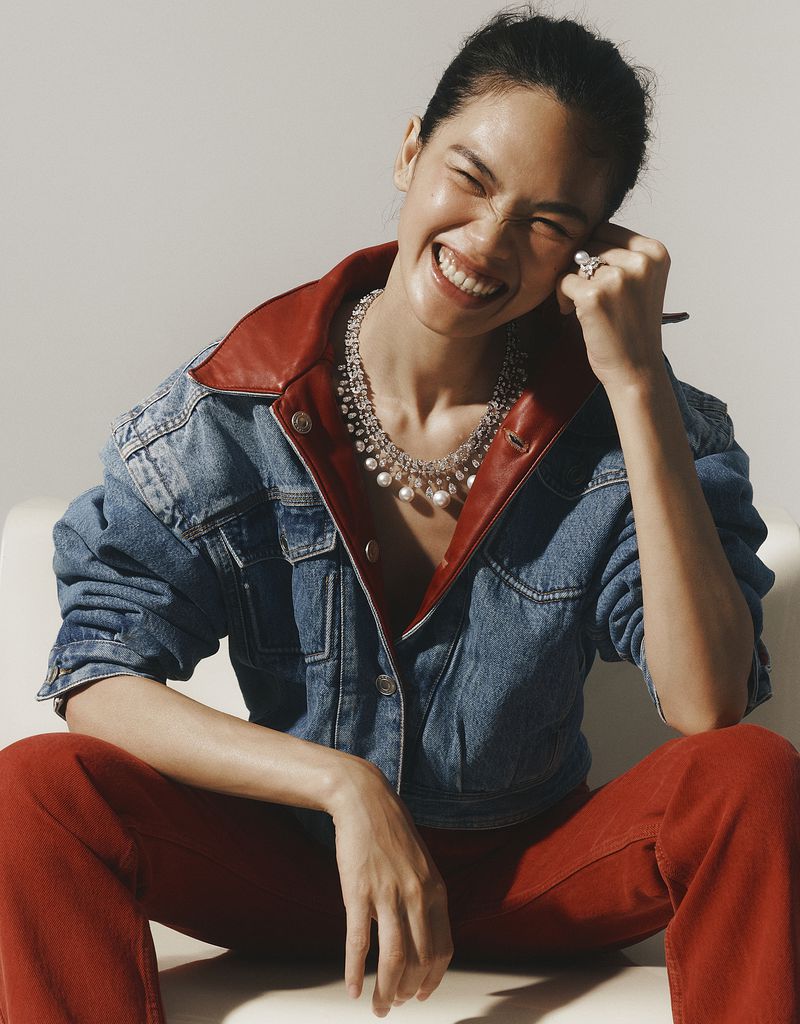Model laughing while wearing a denim jacket, red pants, and statement pearl necklace against a plain background
