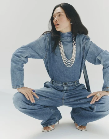Model crouching wearing denim top and jeans, layered pearl necklace, rings, against a plain white background.