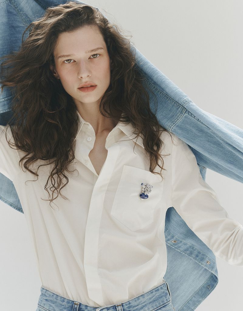 Model with long dark curly hair wearing a white shirt and holding a blue denim jacket in a studio setting.