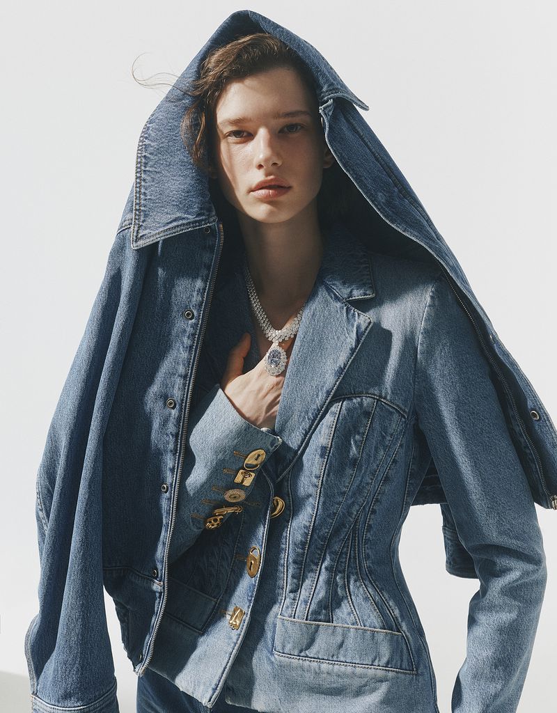 Model wearing a blue denim jacket with a hood and a diamond pendant necklace against a white background