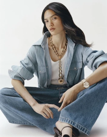 Model seated on the floor wearing a denim shirt, white tank top, jeans, statement chain necklace, and a wristwatch against a white background.