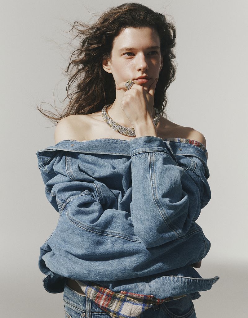 Model wearing a denim jacket and statement jewelry with hand on chin against a plain light background